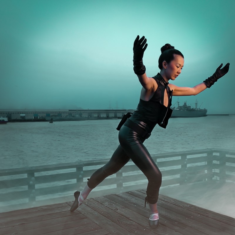 A ballerina clad in cyberpunk black clothes stands, arms raised, waving at ships off the pier.