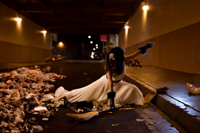 A ballerina in a white dress, wearing a gas mask and gloves is crouching to pick up a broom from the street. She is surrounded by garbage. 