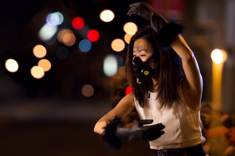 A close up of a ballerina wearing a gas mask, black rubber gloves and a white dress. She is holding one hand over her head and the other in front of her chest. In the distance coloured street lights are blurred. 