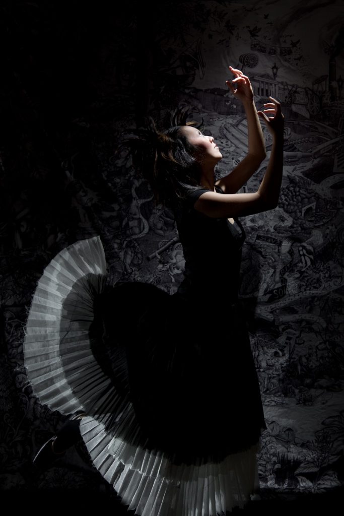A ballerina in a black long dress with white ruffled slip is falling in front of a black and white tapestry. The tapestry shows a world in which coal has no bounds. The ballerina has her hands in the air. She is wearing pigtails and has doll-like black makeup.
