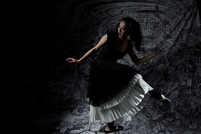 A ballerina in a black long dress with white ruffled slip is sneaking in front of a black and white tapestry. The tapestry shows a world in which coal has no bounds. To the left, a dark shade encroaches. 