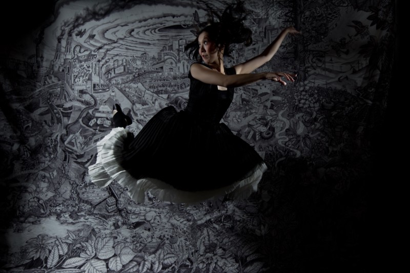 A ballerina in a black long dress with white ruffled slip has jumped magnificently in front of a black and white tapestry. Her hands are both off to the right, while she looks off to the left, frightened. The tapestry shows a world in which coal has no bounds. 