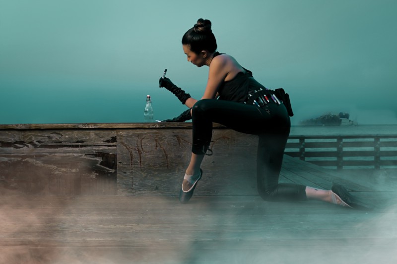 A ballerina clad in black cyberpunk clothes, writes a note, while crouching on the pier. 