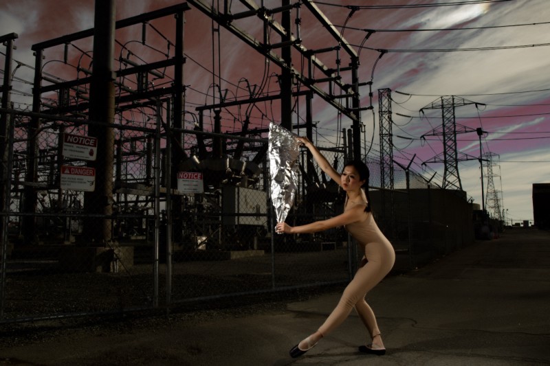 A ballerina in a body suit that matches her flesh tone stands with aluminum foil spread between her hands. She is standing in front of a large electric line grid. The skies are pink, maroons and greys. 
