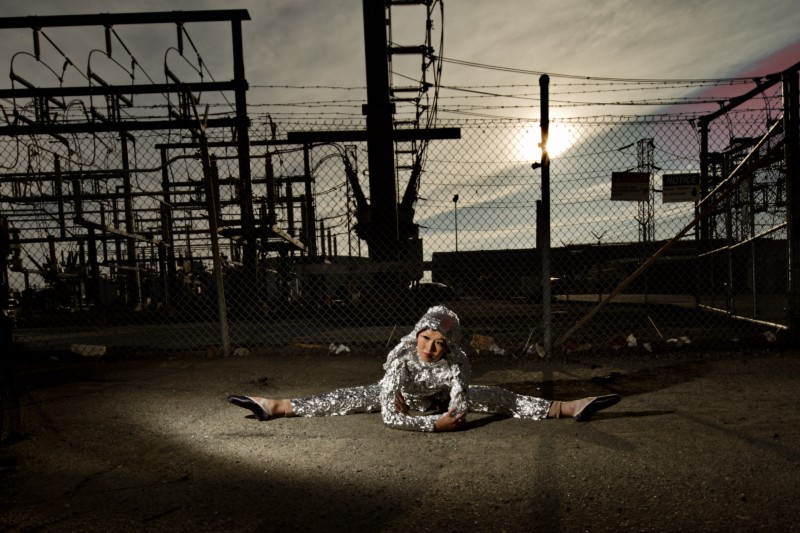 A ballerina in a body suit that matches her flesh tone is laying on her front, doing the splits in front of a fence. She has a full outfit made of aluminum foil. She is in front of a large electric line grid. The skies are yellow, blue and grey. You can see the sun is going down, covered by clouds. 
