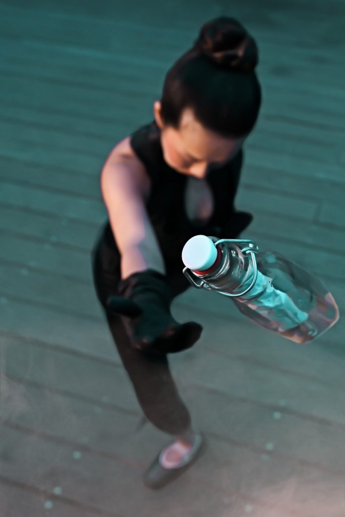 A ballerina stands, head down with her hand up. A bottle with a note inside is in the front of the frame. She has thrown it into the air. 