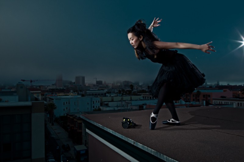 Ballerina in a flowing black dress stands looking over the side of a rooftop. A gasmask is on the roof beside her. 
