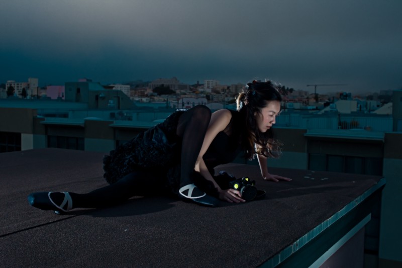 A ballerina in a black flowing dress is crouching on a rooftop, eying something off in the distance.