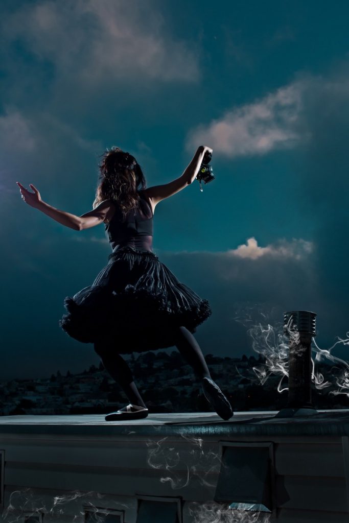 A ballerina holding a gas mask in her hand, stands on a rooftop. A dark cloud is rolling over the hill towards her. A pipe jutting out of the roof has smoke drifting from it. 
