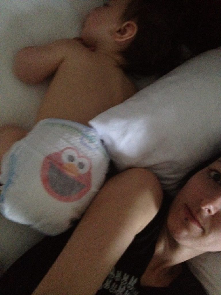 Sleep addled woman with lip piercing laying in bed beside a sleeping baby with it's bum (covered by a diaper)
 beside her head. 